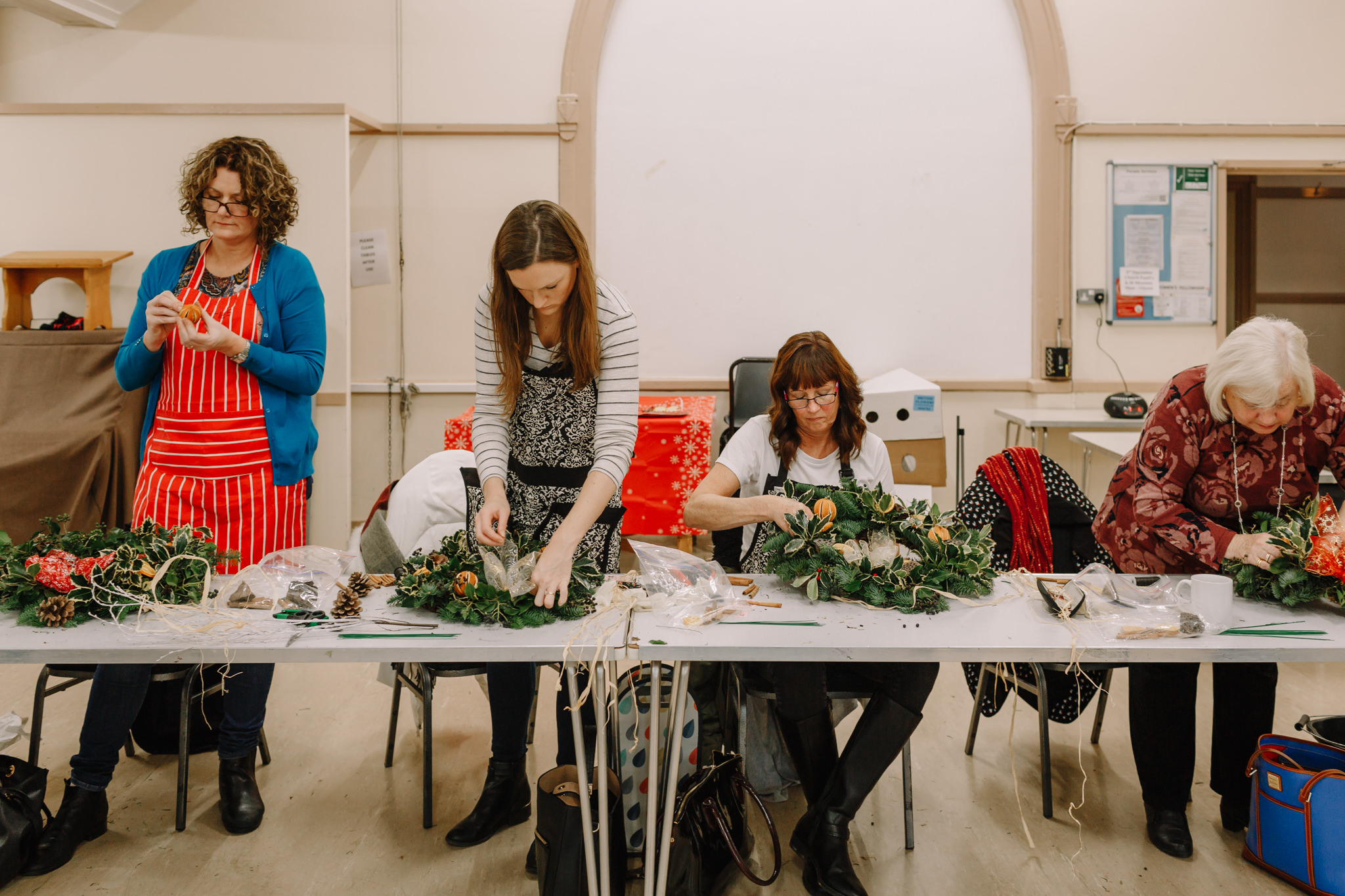 Women sit and stand at table making Christmas wreaths